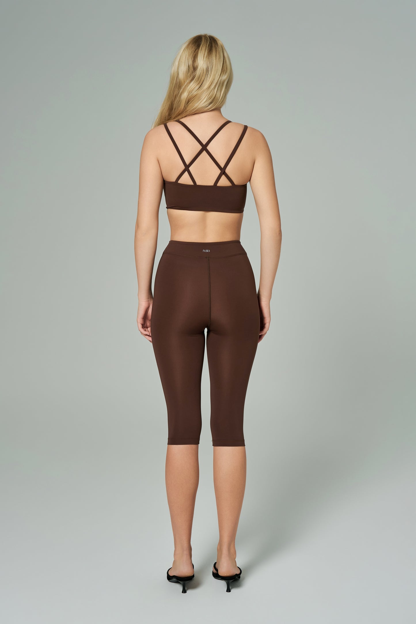 NALA Activewear brown cross-back sports bra and capri leggings set back view; kahverengi çapraz askılı spor sütyen ve diz altı tayt giyen kadın model gri fonda arkadan görünüyor.