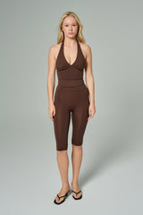 NALA Activewear brown halter biker jumpsuit; kahverengi boyundan bağlamalı kısa paça tulum giyen kadın model gri fonda önden görünüyor.
